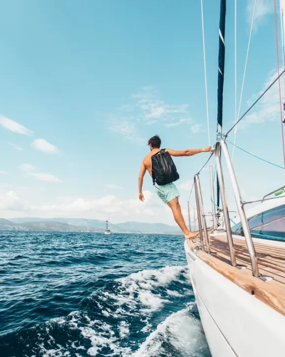 A man with a backpack stands on the edge of a sailboat cruising through vibrant blue waters under a clear sky.